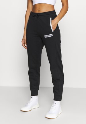 Kobieta ubrana w czarne joggery Calvin Klein z bocznymi kieszeniami oraz białe wysokie trampki, na gładkim tle widać krótki biały top.