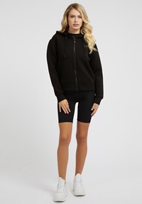 Felpa nera con zip e cappuccio, abbinata a pantaloni corti da motociclista neri. Modella che indossa sneakers bianche, in piedi contro uno sfondo neutro.