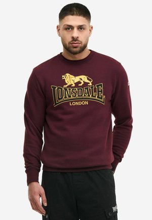 Mees, kes kannab burgundiapunast Lonsdale Londoni pusa kollase lõvi logoga ja musti cargo-pükse, seisab ühevärvilise tausta ees.