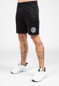 Zwarte sportshorts met een elastische tailleband, met een ronde logo patch aan de linkerkant. G gedragen met witte sneakers.