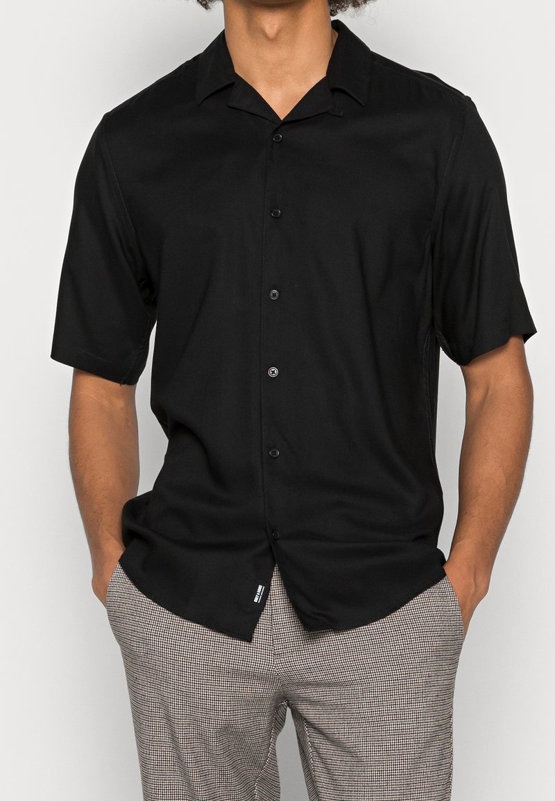 Camisa negra de manga corta con botones, que presenta un cuello extendido y una tela suave. Se lleva con pantalones de patrón a cuadros.