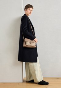 Dlhý čierny kabát cez voľné svetlo modré džínsy, doplnené o čierne dreváky. Hnedá crossbody taška má klopu a kovové detaily.