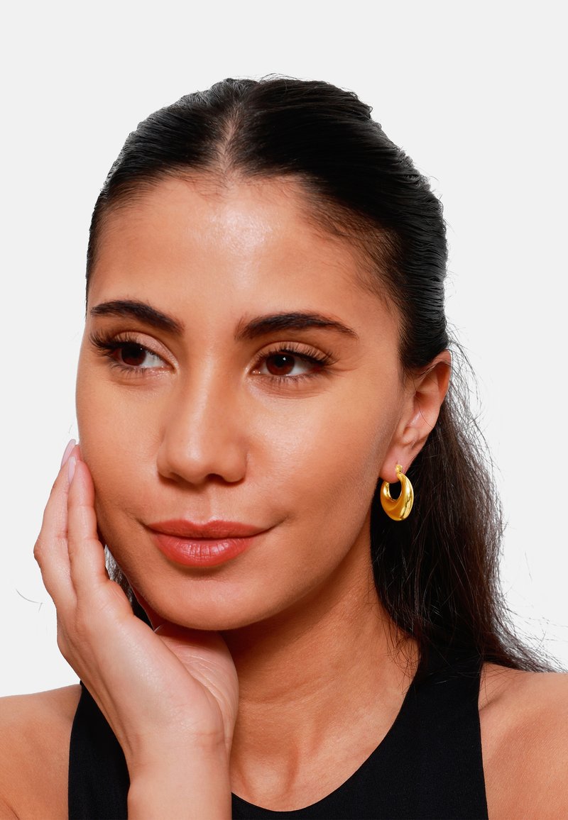 Boucles d'oreilles en or, présentant un design lisse et courbé avec une finition brillante, portées par un modèle aux cheveux foncés, mettant en avant une élégante simplicité.