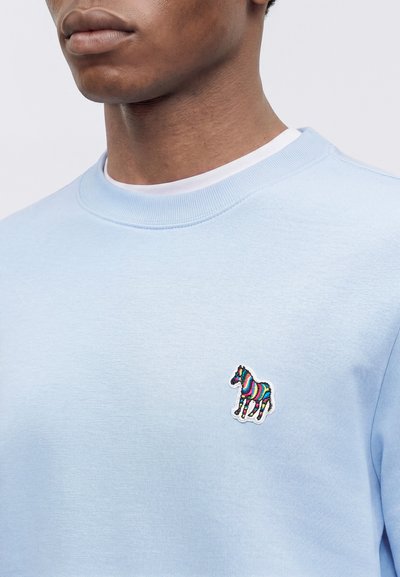 Homme portant un sweat bleu clair avec un patch zèbre rayé multicolore sur la poitrine gauche, superposé à une chemise blanche.