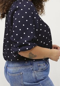 Granatowa bluzka w białe kropki, z długimi rękawami i luźnym krojem. Połączona z klasycznymi niebieskimi dżinsami. Teksturowany materiał z subtelnym połyskiem.