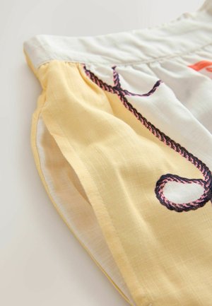 Detailní záběr na látkovou blůzu z žluté bavlny s vyšívaným černým a růžovým provázkovým vzorem podél bočního švu. Lehká a texturovaná.