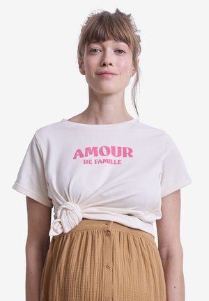 Frau mit hellbraunen Haaren, die ein gebundenes weißes T-Shirt mit pinkem "AMOUR DE FAMILLE"-Schriftzug und einen hoch taillierten braunen Rock mit Knöpfen trägt, schaut nach vorne.
