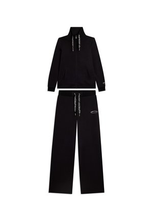 Tuta nera due pezzi con giacca con zip e pantaloni ampi, con lacci bianchi marchiati e piccolo testo bianco su petto e gamba.