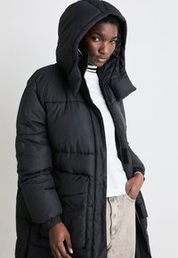 Black puffer coat featuring a hood, zip closure, elastic cuffs, and side pockets. Textured, insulated material with a sleek design.
