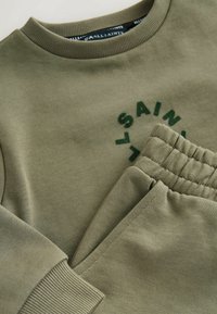 Olivengrüner Strickpullover mit geripptem Ausschnitt und Bündchen, mit gesticktem "ALLSAINTS"-Logo. Passende elastische Hose ist sichtbar.