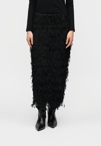 Μαύρη φούστα με κρόσσια από υλικό με υφή, μέχρι το γόνατο, συνδυασμένη με μαύρα μπότες μέχρι τον αστράγαλο με μυτερή μύτη. Ο σχεδιασμός περιλαμβάνει στρωματοποιημένα κρόσσια.