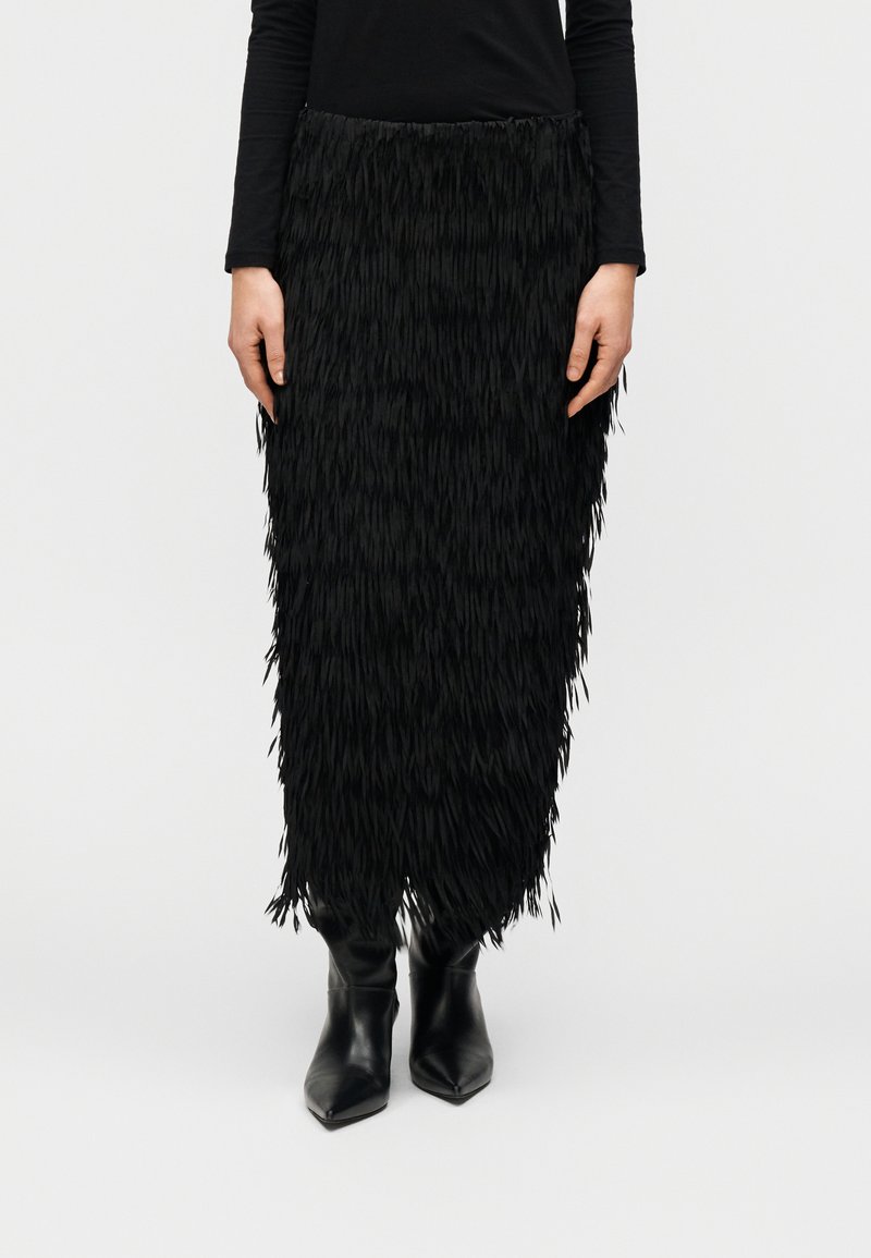 Μαύρη φούστα με κρόσσια από υλικό με υφή, μέχρι το γόνατο, συνδυασμένη με μαύρα μπότες μέχρι τον αστράγαλο με μυτερή μύτη. Ο σχεδιασμός περιλαμβάνει στρωματοποιημένα κρόσσια.