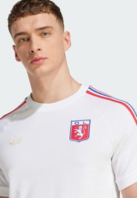 Giovane uomo con capelli ricci corti che indossa una maglietta sportiva bianca con strisce rosse e blu e un emblema di leone sul petto.