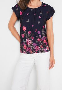 Marinblå blus med blommönster, korta fladdrande ärmar, livfulla rosa och lila blommor samt en rynkad halsringning; tyget verkar vara lätt och slätt.