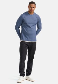 Lichtblauw geribbeld shirt met lange mouwen en een ronde hals, gedragen over een wit shirt, gecombineerd met donkere spijkerbroek en witte sneakers.