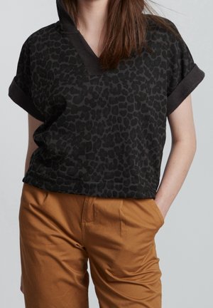 Femme portant un haut noir à manches courtes imprimé léopard et un pantalon beige, une main dans la poche, sur un fond uni.