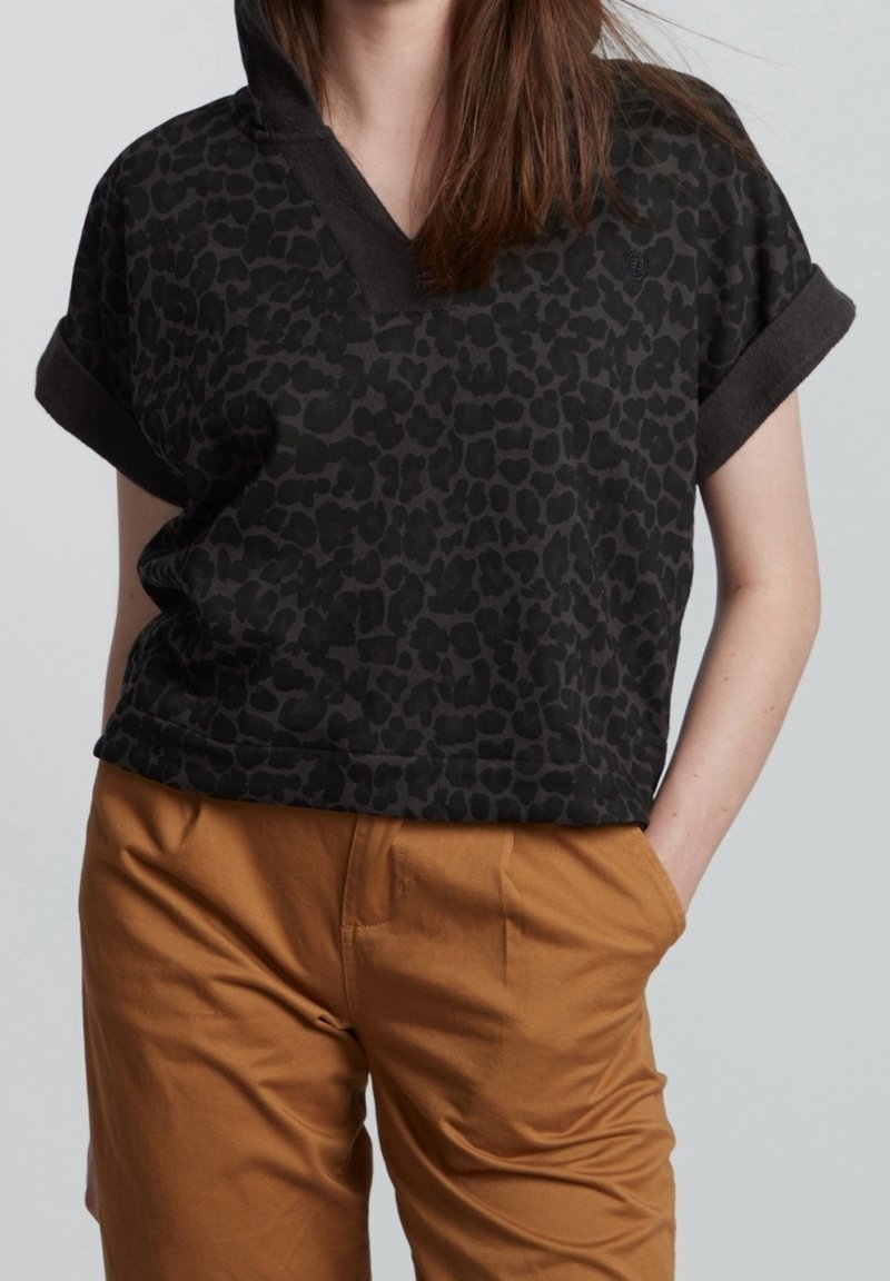 Femme portant un haut noir à manches courtes imprimé léopard et un pantalon beige, une main dans la poche, sur un fond uni.