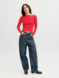 Kvinne som står med hendene i lommer, iført rød langermet topp, løse blå jeans og svarte sko med firkantet tå, mot en ensfarget bakgrunn.