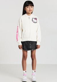 Felpa con zip di colore crema con grafica di Hello Kitty, dettagli rosa sulle maniche, abbinata a una gonna di jeans nera e calzini con lo stesso personaggio.