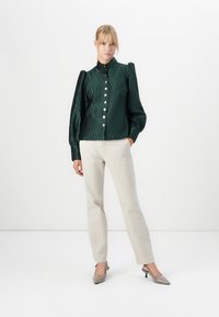 Kvinne iført en teksturert mørkegrønn bluse med knapper og puffermer, beige bukser med rett bein, og spisse taupe hæler, stående mot hvit bakgrunn.