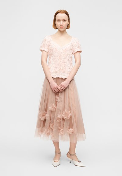 Γυναίκα όρθια με τα χέρια σταυρωμένα, φορώντας ένα ανοιχτό ροζ τοπ με υφή λουλουδιών, διάφανο μίντι φόρεμα με υφασμάτινα λουλούδια και λευκά παπούτσια με χαμηλό τακούνι.