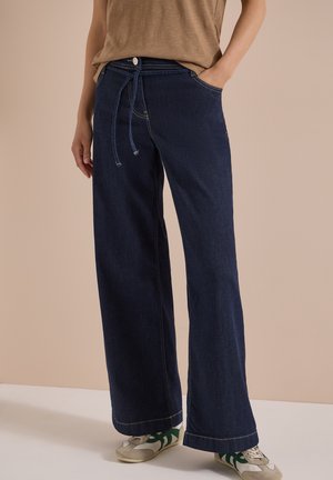 Personne portant un jean bleu foncé à jambes larges avec une taille à cordon, un haut beige et des baskets blanches avec des bandes vertes.