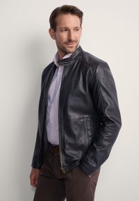 Czarna skórzana kurtka z kołnierzem stojącym, wyposażona w przedni zamek błyskawiczny i dwie boczne kieszenie. Nosi się ją na jasną koszulę w kratę.