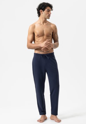 Hemdloser Mann mit Tattoos, der dunkelblaue Jogginghosen trägt, barfuß vor einem schlichten weißen Hintergrund steht und zur Seite schaut.