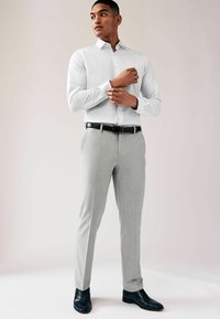 Camisa blanca de manga larga con cuello extendido, combinada con pantalones grises claros y zapatos de vestir negros. Presenta un tejido suave y un ajuste entallado.
