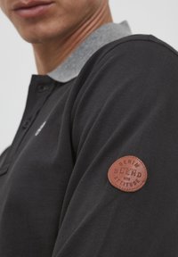 Svart polo skjorte med grå krage, teksturert stoff, med et brunt lærlogo-lapp på ermet som sier "DENIM BLEND med ATTITUDE."