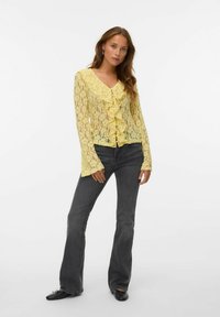 Camicia gialla in pizzo con fronte a volant, maniche lunghe e scollo a V. Indossata con jeans a zampa d'elefante grigio scuro e scarpe nere. Sfondo pulito.