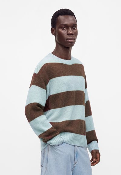 Jeune homme aux cheveux courts portant un pull ample à manches longues avec de larges rayures horizontales bleues et marron et un jean bleu clair.
