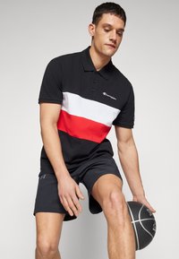 Schwarzes Polo mit weißen und roten horizontalen Streifen, aus atmungsaktivem Stoff. Getragen mit schwarzen Athletics-Shorts und während er einen Basketball hält.