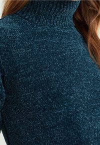 Smaragdgrön polotröja med en texturerad, ribbad design. Tyget ser mjukt och varmt ut, vilket framhäver den varma, plyschiga ytan.