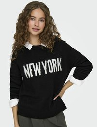 Maglione nero con testo bianco "NEW YORK", scollatura a girocollo e maniche lunghe con polsini arrotolati sopra una camicia bianca con colletto. Texture morbida.