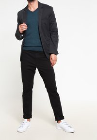 Mężczyzna w turkusowym swetrze w serek, czarnym żakiecie, czarnych spodniacach podwiniętych i białych sneakersach stoi na tle białej ściany.