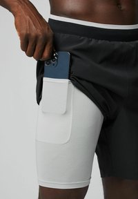 Schwarze Sportshorts mit einer weißen Mesh-Tasche, in der ein Smartphone gehalten wird. Der elastische Bund hat eine glatte Textur.