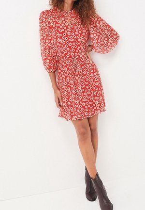 Robe fleurie rouge avec manches bouffantes, taille froncée et décolleté en trou de serrure, ornée de marguerites blanches et d'une ceinture nouée. Bottines marron.