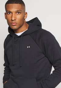 Sudadera negra con capucha, bolsillo frontal, cordones y un pequeño logo blanco en el pecho. Confeccionada en un tejido suave y texturado.