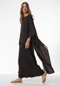 Schwarzes, fließendes Tunika-Oberteil mit weiten Ärmeln und passendem langen Rock. Leichter Stoff, glatte Textur, mit gestuftem Design. Goldene Sandalen.