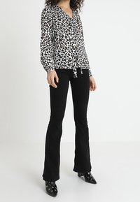 Vit blus med svart och brun leopardmönster, långa ärmar, v-ringning, knuten framtill, i kombination med svarta utsvängda byxor och spetsiga svarta skor.