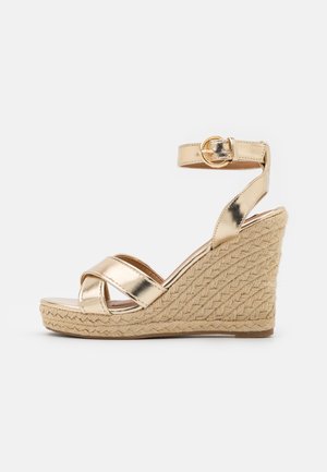 Sandale compensée dorée métallique avec talon en jute tressé, bride à la cheville avec boucle, et lanières croisées décoratives sur l'avant-pied.