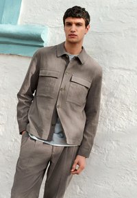 Chaqueta de lino gris con dos bolsillos en el pecho, frente con botones y cuello. Combinada con pantalones holgados a juego y una camiseta azul claro.