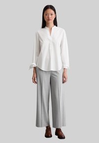 Camicia bianca a maniche lunghe con colletto cinese e orlo leggermente curvo, abbinata a pantaloni larghi grigi chiari e scarpe stringate marroni.