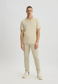 Helles beige Polohemd mit gelben Akzenten, kurzen Ärmeln und Kragen, kombiniert mit passenden beigen Hosen und weißen Sneakers.