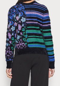 Mångfärgad stickad tröja med en fluffig textur, som har blommönster i lila och blått, samt randiga sektioner i grönt och svart.