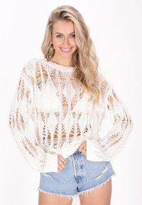 Maglione bianco lavorato a maglia con motivo a pizzo aperto, scollo rotondo e maniche lunghe, abbinato a pantaloncini di jeans chiari con bordi sfilacciati.