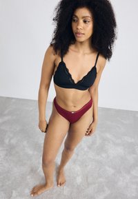 Musta pitsireunainen bralette ja burgundivärinen stringi, molemmat valmistettu sileästä kankaasta. Malli poseeraa vaaleanharmaalla lattialla yksinkertaista taustaa vasten.