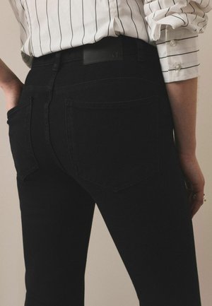 Person wearing black high-waisted jeans and a white long-sleeve shirt with black vertical stripes, hand in back pocket.