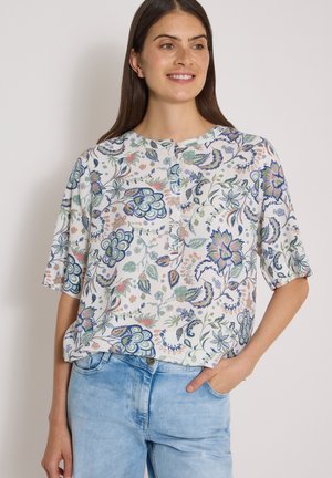 Femme portant un chemisier blanc à manches courtes avec des motifs floraux colorés et un jean bleu clair, souriante avec une main dans la poche.